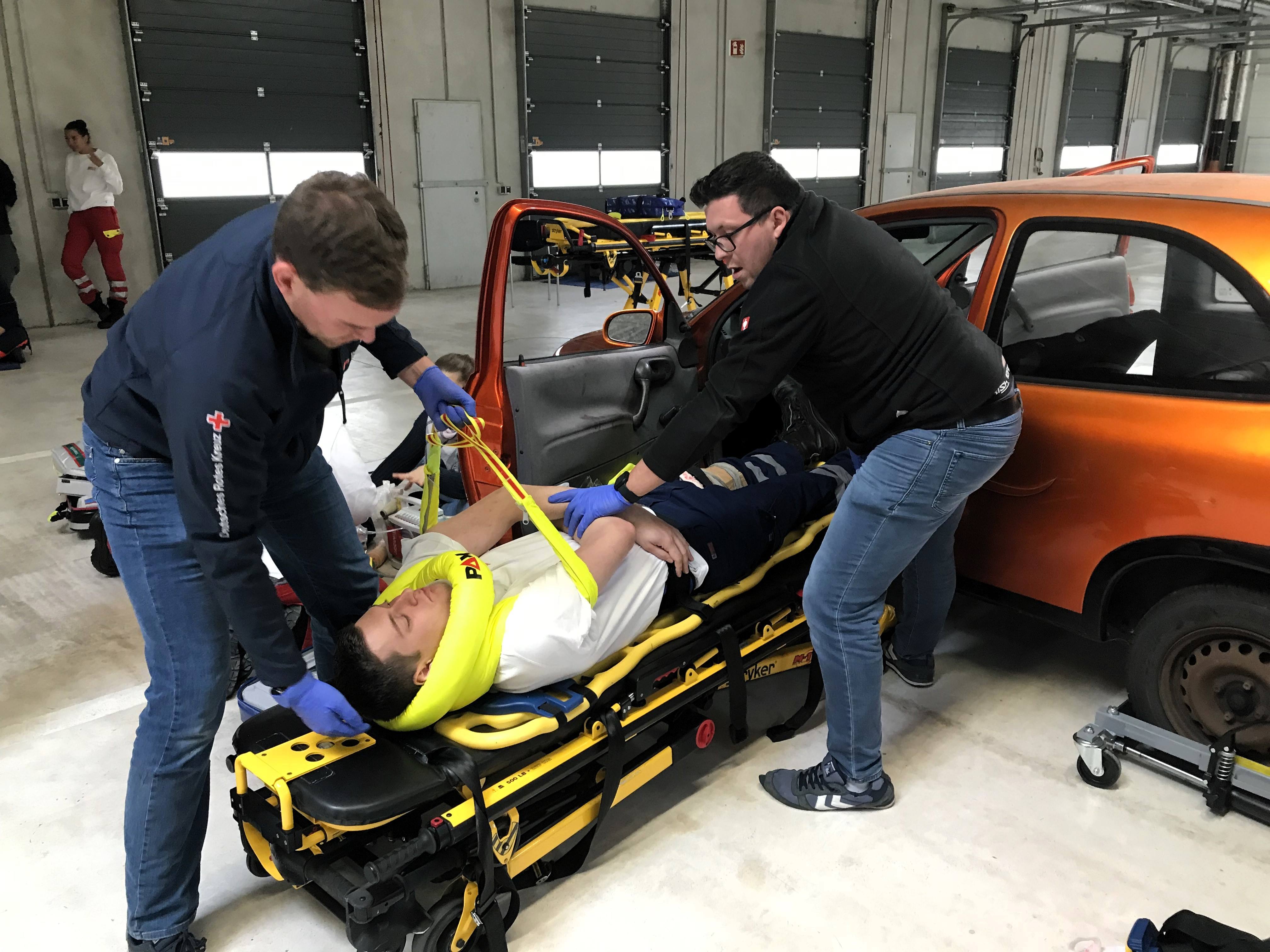 Ein Mann wird von zwei Sanitätern bei einem simulierten Autounfall mit einer Trage aus dem Auto gehoben. Sein Nacken wird durch eine Nackenkrause gestützt.