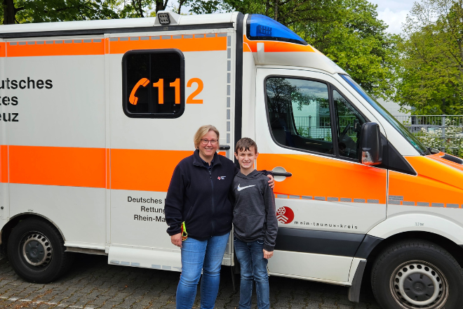 Eine Mitarbeiterin des DRK steht mit dem Besucher vom Zukunftstag vor einem Rettungswagen.