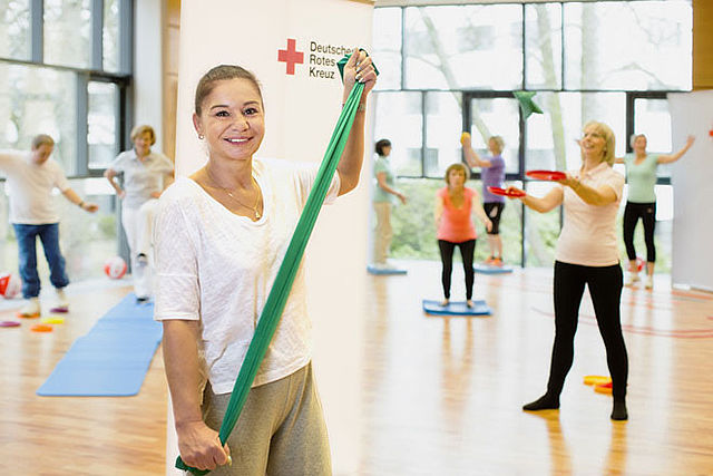 Eine Dame steht mit einem grünen, gespannten Gymnastikband zwischen den Händen im Vordergrund. Im Hintergrund sind verschwommen andere Personen zu sehen, die verschiedene Sport- bzw. Gymnastikübungen machen.