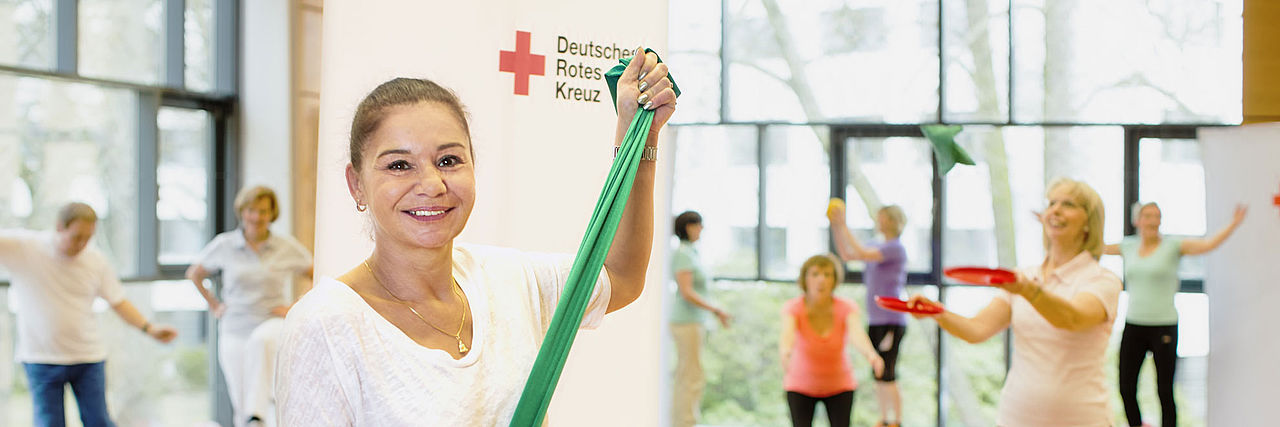 Eine Dame steht mit einem grünen, gespannten Gymnastikband zwischen den Händen im Vordergrund. Im Hintergrund sind verschwommen andere Personen zu sehen, die verschiedene Sport- bzw. Gymnastikübungen machen.