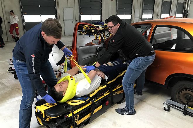 Ein Mann wird von zwei Sanitätern bei einem simulierten Autounfall mit einer Trage aus dem Auto gehoben. Sein Nacken wird durch eine Nackenkrause gestützt.
