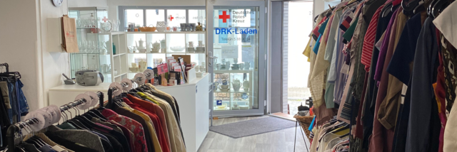 Innenansicht des Kleiderladens mit Kleiderständern. Eine große Fensterfront ist mit Geschirr geschmückt. Die Tür steht offen.