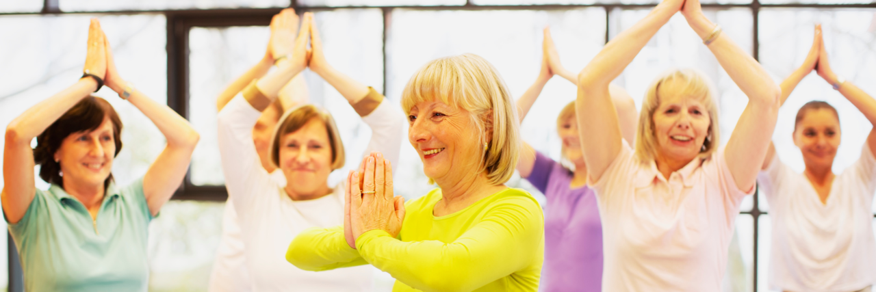 Mehrere Damen machen Yoga. Dabei steht eine Frau im gelben Oberteil vorne und zeigt den anderen die Posen.